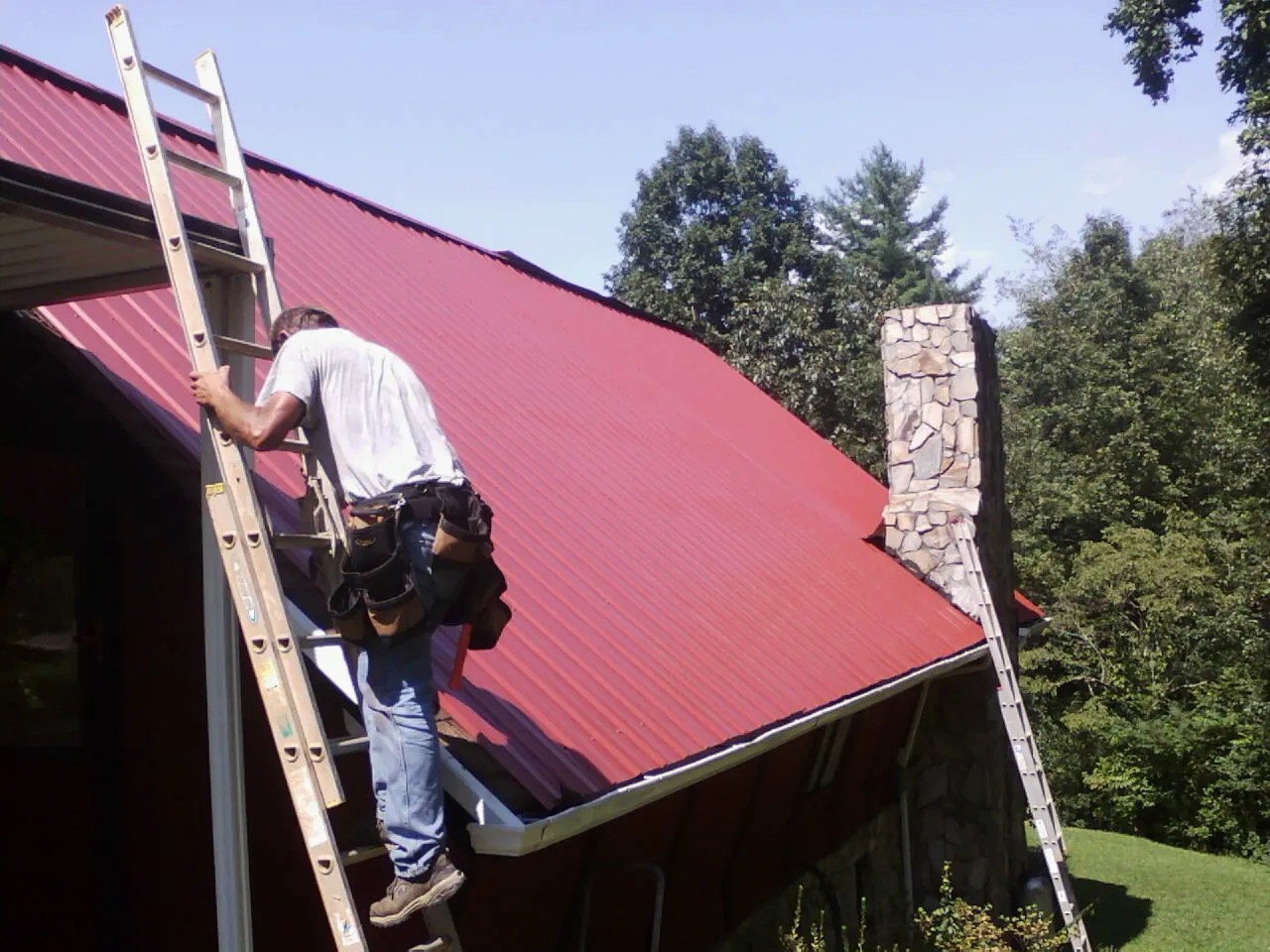 Roofer on a ladder beside a red metal roof and stone chimney — Soffit & Fascia Repair by Roofing Brothers in Winslow